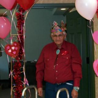 An elderly man wearing a festive crown stands amidst heart-themed decorations and balloons, symbolizing a joyful celebration.