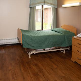 A hospital patient room featuring a bed with green bedding, wooden flooring, and natural light coming in through the window.