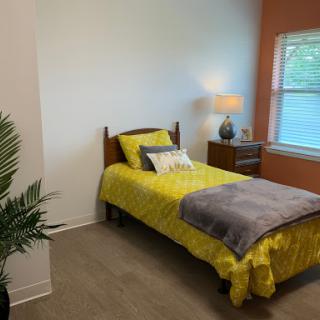 A cozy bedroom featuring a single bed with vibrant yellow bedding, complemented by a decorative lamp and a touch of greenery.