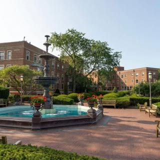 A peaceful park scene with a central fountain, surrounded by benches and greenery, set against the backdrop of brick buildings on a sunny day.