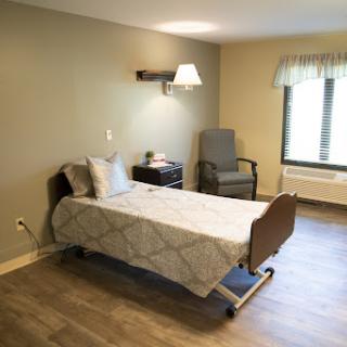 A clean and simple hospital patient room featuring a bed, a chair, and natural lighting.