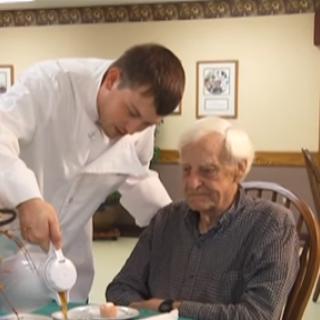 A caregiver pours tea for an elderly man, showcasing a caring interaction in a communal dining setting.