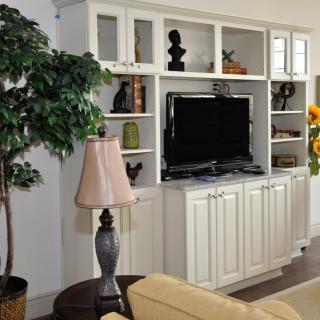 A cozy living room with a white entertainment center, featuring a television, decorative items on shelves, and a nearby table lamp.