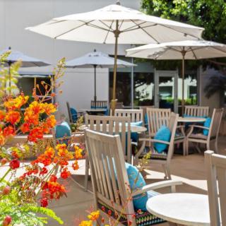 A sunny patio with wooden tables, chairs, and umbrellas surrounded by vibrant flowers.