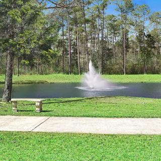 A serene park scene with a fountain in a pond, surrounded by trees, grass, and a bench.