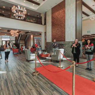 A spacious hotel lobby featuring a red carpet walkway, elegant chandeliers, and guests mingling in the reception area.