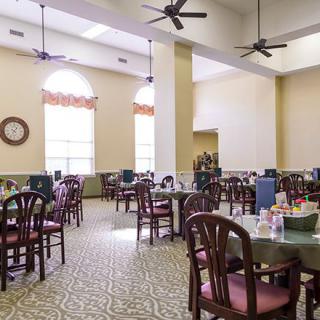 An empty dining room with multiple tables and chairs, featuring large windows and ceiling fans.
