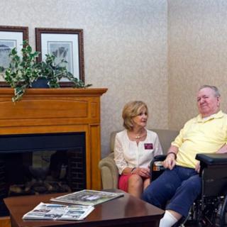 Two individuals sitting in a lounge area, one in a wheelchair, engaging in conversation near a fireplace.