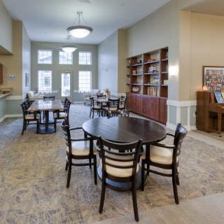 A well-furnished library room with tables and chairs, featuring bookshelves and ample natural light.