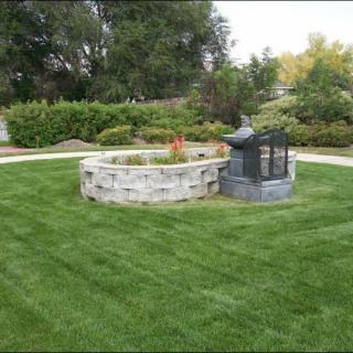 A well-maintained lawn with a circular stone fire pit in the center of a landscaped garden.
