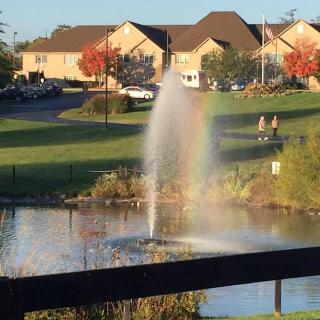 A serene park scene with a fountain creating a rainbow effect over a lake, surrounded by suburban homes and autumn trees.