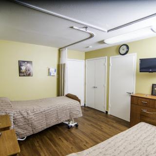 A spacious hospital room featuring two patient beds, a clock on the wall, and a warm yellow color scheme.