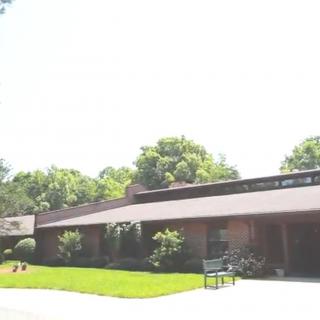 A one-story building surrounded by trees and a well-kept lawn, with a bench in front.