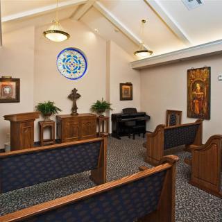 A small chapel interior with pews, an altar, and a piano, featuring a stained glass window.
