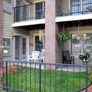 A cozy apartment balcony with outdoor furniture, hanging and potted plants, bordered by a black fence and a small green lawn.