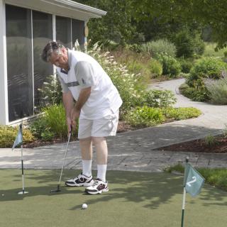A person practices putting on an outdoor green surrounded by a garden.