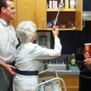 An elderly woman receives assistance in the kitchen, using a cane to reach items on a high shelf.