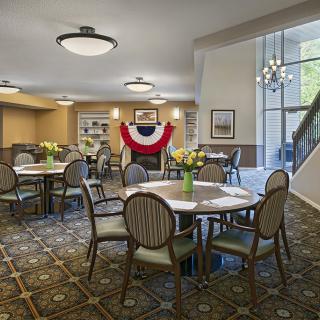 A well-lit dining area featuring multiple round tables and chairs, decorated with a patriotic banner and floral arrangements.