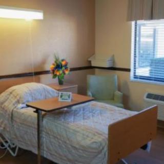 A hospital room with a single bed, side table, chair, and flowers by the window.