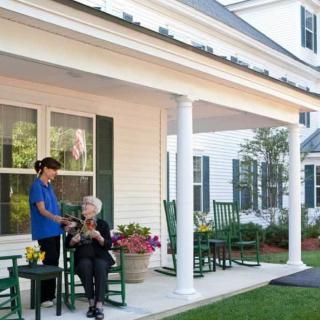A caregiver and a senior person talking on a house porch surrounded by greenery.
