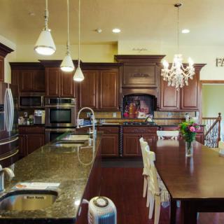 A modern kitchen with wooden cabinetry and granite countertops, featuring a large island and dining table under elegant lighting fixtures.