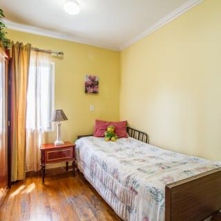A cozy bedroom with a single bed and warm yellow walls, featuring wooden flooring and a window with sheer curtains.