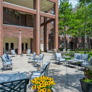 An inviting outdoor patio area with comfortable seating, surrounded by sunflowers and trees, adjacent to a brick building.