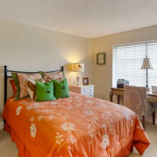 A cozy bedroom with an orange bedspread and green pillows, featuring a lamp on a nightstand and a desk by the window.