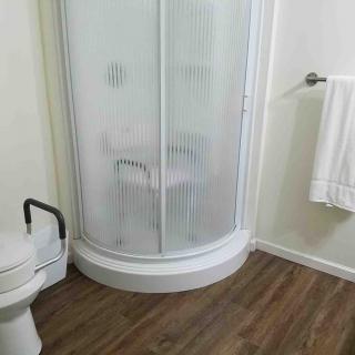 A bathroom featuring a corner shower with frosted glass, a toilet with a handrail, and a towel rack on a wooden floor.
