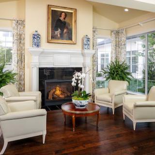 A cozy living room with a fireplace, surrounded by armchairs and a coffee table, decorated with plants and a portrait above the mantel.