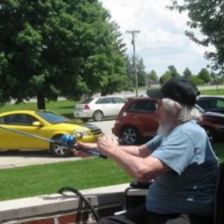 An elderly person sits outside on a sunny day, pointing a toy gun while cars pass by.