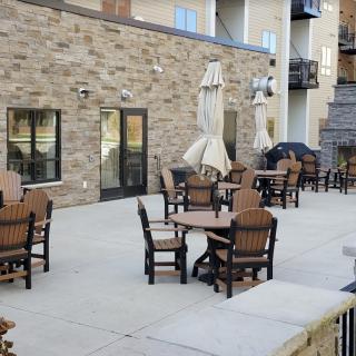 An outdoor patio area with tables and chairs, set against a brick wall, featuring closed umbrellas.