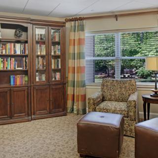A cozy living room with a bookshelf, armchair, and ottomans near a window.