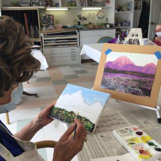 An artist working on a landscape painting in an art studio, with a completed mountain scene on display.