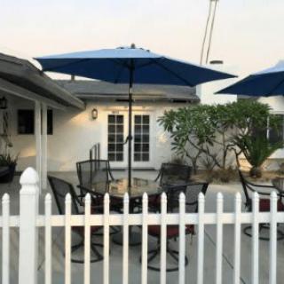 A cozy backyard patio area with tables, chairs, blue umbrellas, and a white picket fence.