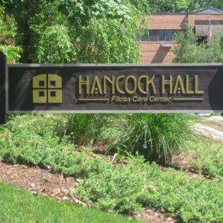A sign for Hancock Hall in front of a landscaped area with greenery and a building in the background.