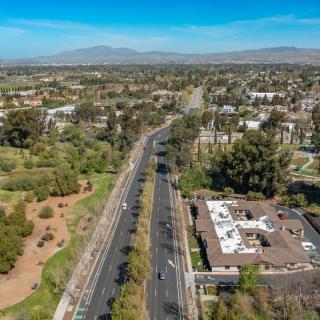 Aerial view of an urban landscape featuring a road lined with trees, buildings, and mountains in the background under a clear blue sky.