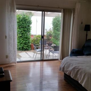 A cozy bedroom with wooden flooring opens up to a patio through sliding glass doors, offering a lush garden view.