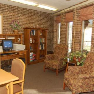 A cozy office space with a computer, bookshelf, chairs, and large windows.