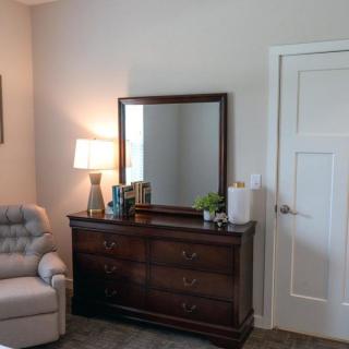 A cozy bedroom corner featuring a dresser with a mirror, a lamp, and an upholstered chair.