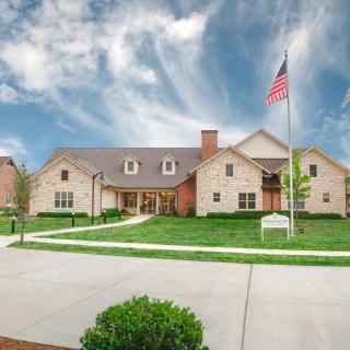 A large residential or institutional building with an American flag on a pole, surrounded by a well-maintained lawn and a driveway under a cloudy sky.