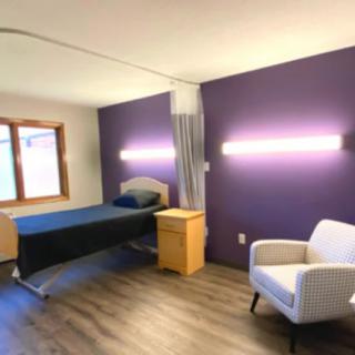 A minimalist bedroom featuring a single bed with a purple accent wall, wooden flooring, and soft lighting.