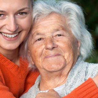 A young woman embraces an elderly woman, both smiling warmly outdoors.