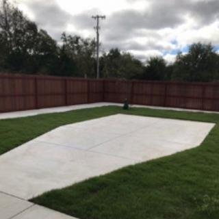 A backyard with a concrete patio surrounded by grass and a wooden fence under a cloudy sky.