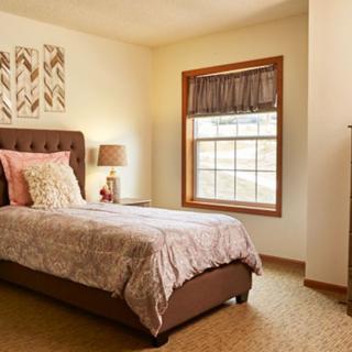 A cozy bedroom with a single upholstered bed, a side table with a lamp, and a window framed by wooden trim.