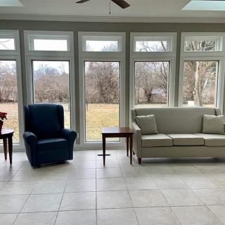 A bright living room with large windows overlooking the outdoors, featuring a sofa, armchair, and small tables.