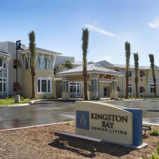 The entrance of Kingston Bay Senior Living facility with a welcoming sign and palm trees lining the pathway.