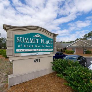 A sign for Summit Place of North Myrtle Beach, with a building and cars in the background under a partly cloudy sky.