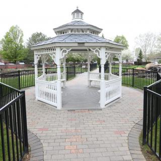 A white gazebo situated on a brick pathway in a park setting, surrounded by greenery.