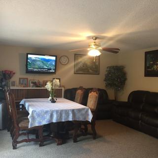 A cozy living room featuring a dining table, sofas, a television on the wall, and a ceiling fan.